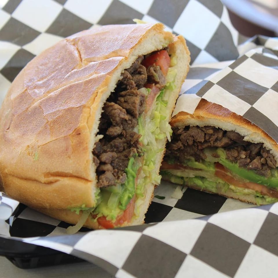 A Boss Man Tacos Burritos & More Mexican torta, cut in half, filled with meat, lettuce, avocado, and tomato.