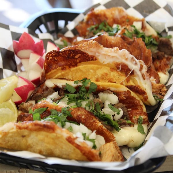 Close-up of cheesy Boss Man Tacos with meat, onions, cilantro, radishes, and lime wedges.