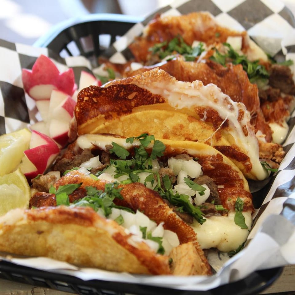 Close-up of cheesy Boss Man Tacos with meat, onions, cilantro, radishes, and lime wedges.