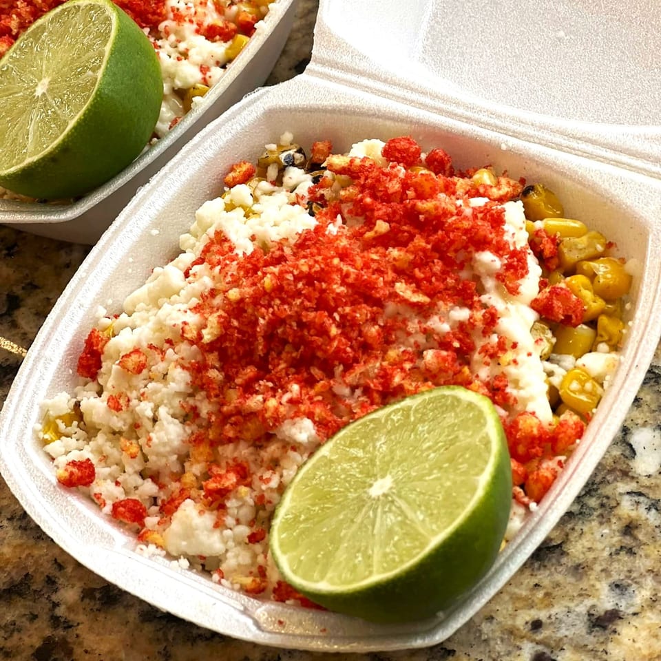 Mexican street corn (Esquites) with cheese, chili powder, and a lime wedge in a to-go container.